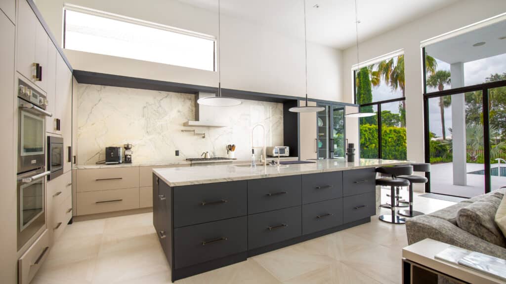Modern kitchen with a large island, marble countertops, wall-mounted ovens, pendant lights, floor-to-ceiling windows, and an outdoor view with palm trees.