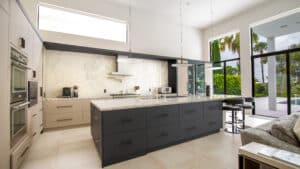 Modern kitchen with a large island, marble countertops, wall-mounted ovens, pendant lights, floor-to-ceiling windows, and an outdoor view with palm trees.