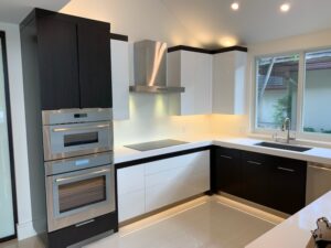 Modern kitchen with black and white cabinets, built-in oven, stovetop with hood, and a sink under a window. Bright lighting and minimalistic design.