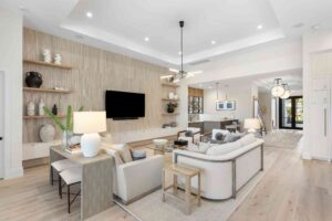 Modern living room with neutral tones, featuring a sofa, armchairs, lamps, and a wall-mounted TV. Custom cabinets showcase decorative items on the shelving while offering full access cabinetry. Enjoy the seamless view into the dining area and entrance hallway.