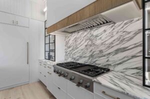 Modern kitchen with marble backsplash, stainless steel gas stovetop, and custom white cabinets. Vertical handles adorn the high-end cabinetry, while a built-in oven is visible on the left.